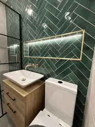 white ceramic basin on oak vanity with brass wall-mounted tap and toilet against green herringbone tiled bathroom wall with illuminated recessed mirror