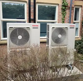 Two white outdoor AC units beside a brick building, with windows and overgrown shrubs in front.